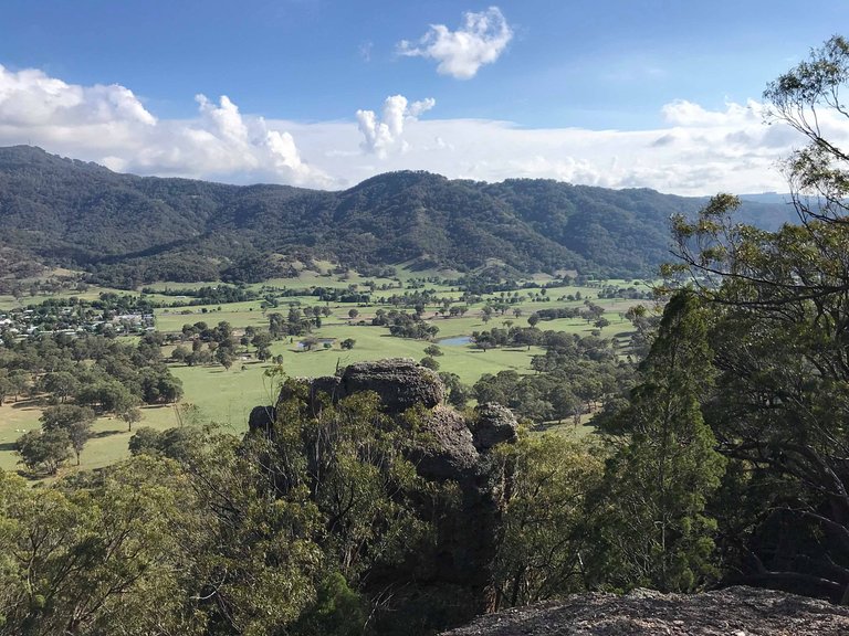A panoramic view of rolling hills and green fields under a partly cloudy sky, with rocky formations in the foreground.
